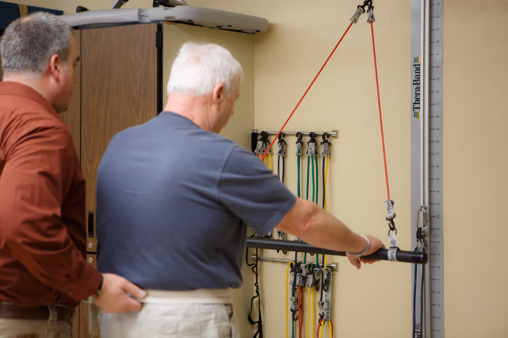 An elderly man performs a resistance band exercise using a TheraBand pulley system mounted on a wall, assisted by a caregiver or therapist standing beside him in a rehabilitation or therapy room.