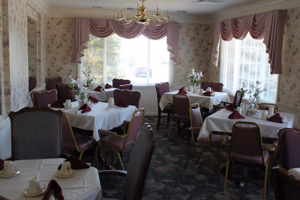 A dining room with multiple tables covered with white tablecloths, each set with cups, saucers, and folded burgundy napkins. The room has floral wallpaper, large windows with pink valances, and floral centerpieces on the tables.