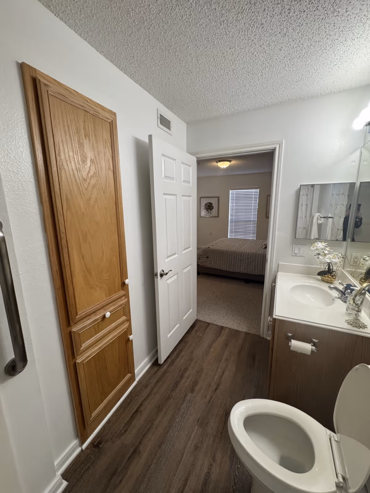 Interior view of a bathroom with a wooden cabinet, white toilet, and sink with a mirror above it. The bathroom has wood-style flooring and white walls. The door to the bathroom is open, revealing a bedroom with a bed, window with blinds, and framed artwork on the walls.