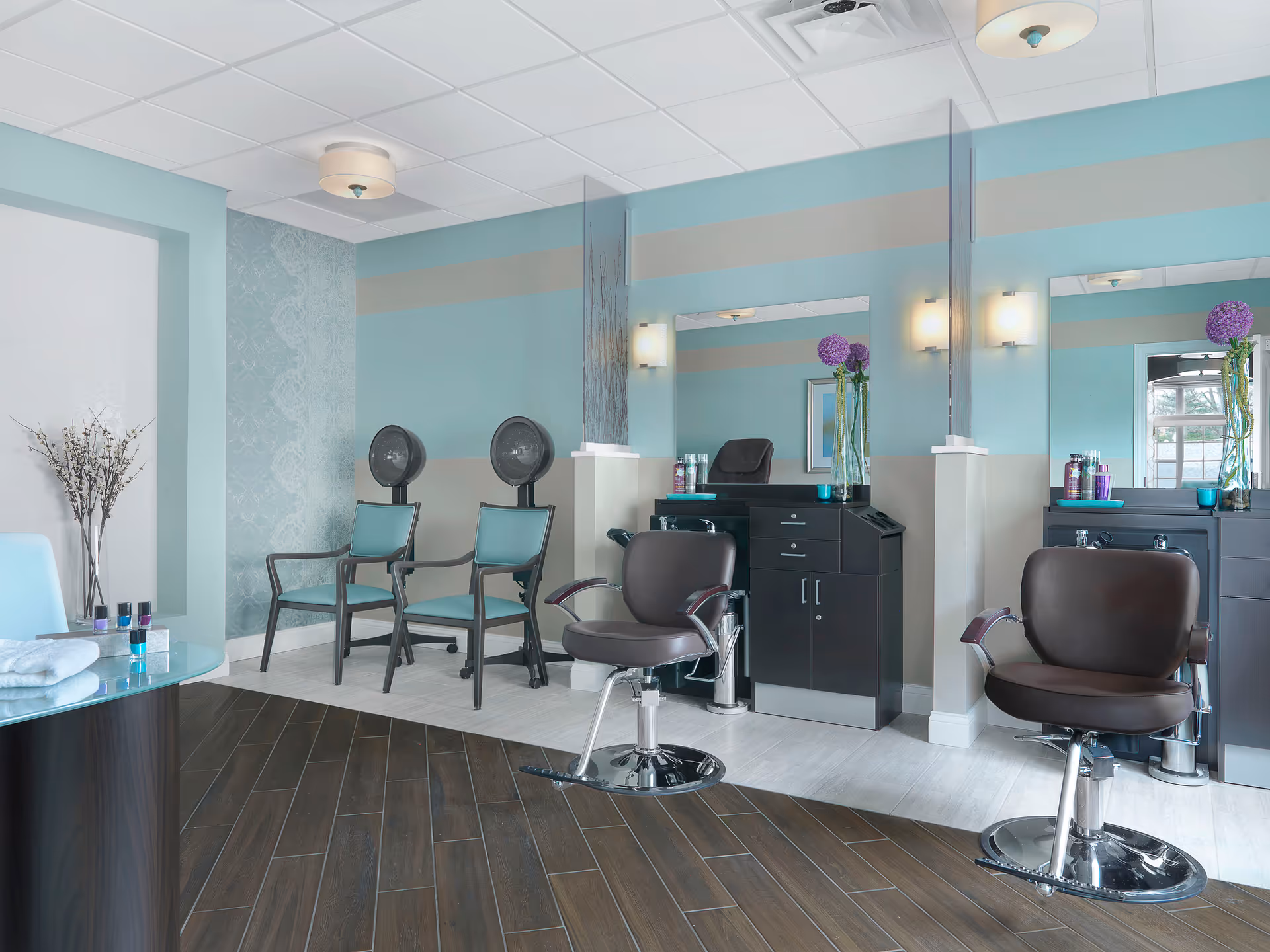 A modern salon area with two brown styling chairs in front of mirrors and black cabinets. Two teal chairs with hair dryers are positioned against a light blue and beige wall. A small table with towels and nail polish is visible on the left side, and decorative flowers are placed on the cabinets and in a vase on the floor.