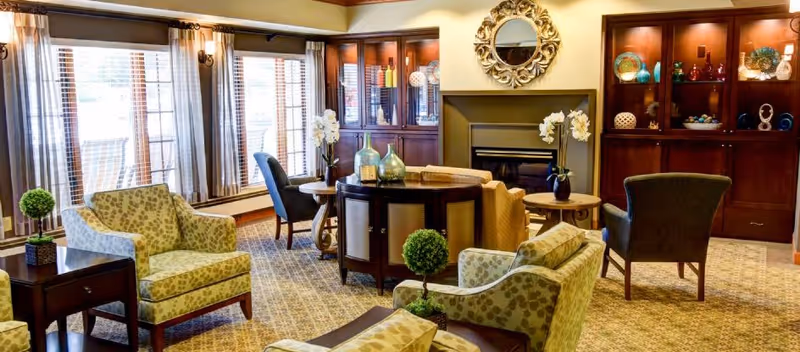 Well-appointed common living room with upholstered armchairs, a round central table, a fireplace with decorative mirror, built-in display cabinets, and large windows with curtains.