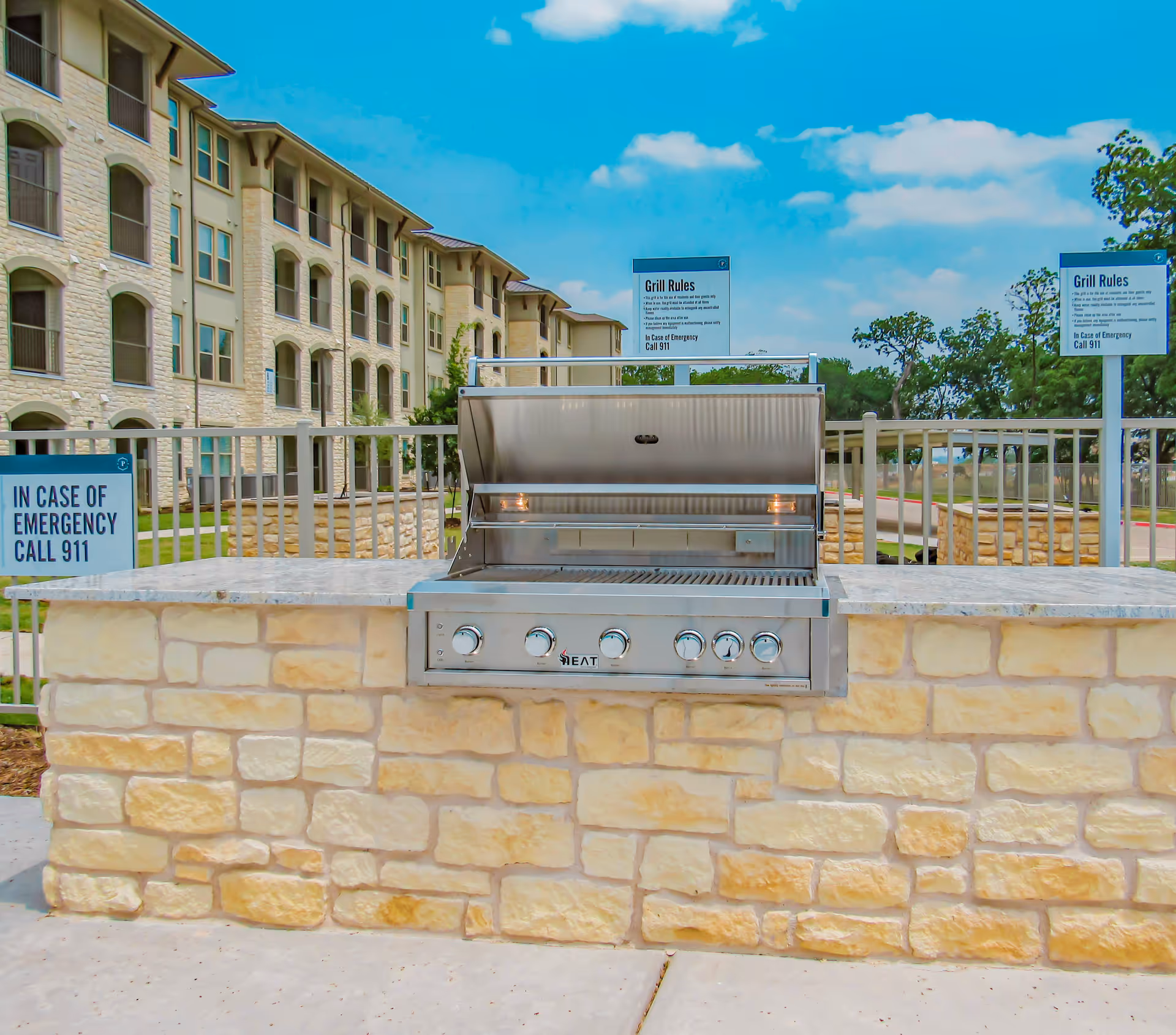 Stainless steel outdoor grill set into a stone counter with a multi-story senior living building and signage in the background.