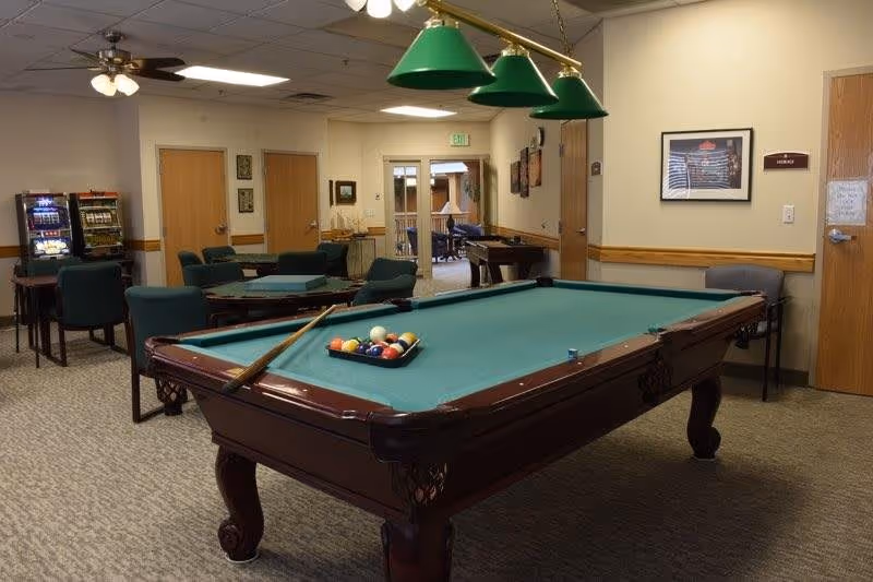 A recreational room with a pool table in the foreground, complete with billiard balls and a cue stick. The room has green cushioned chairs around tables, ceiling fans, and a slot machine in the back corner. There is a doorway leading to an outdoor patio area.