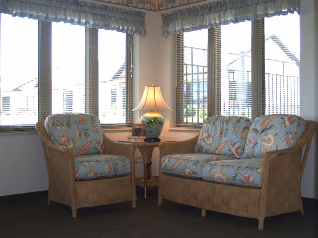A cozy corner seating area with a wicker armchair and loveseat, both upholstered in floral patterned fabric. Between them is a small wooden side table with a decorative lamp. The background features several large windows with blinds and valances, letting in natural light.