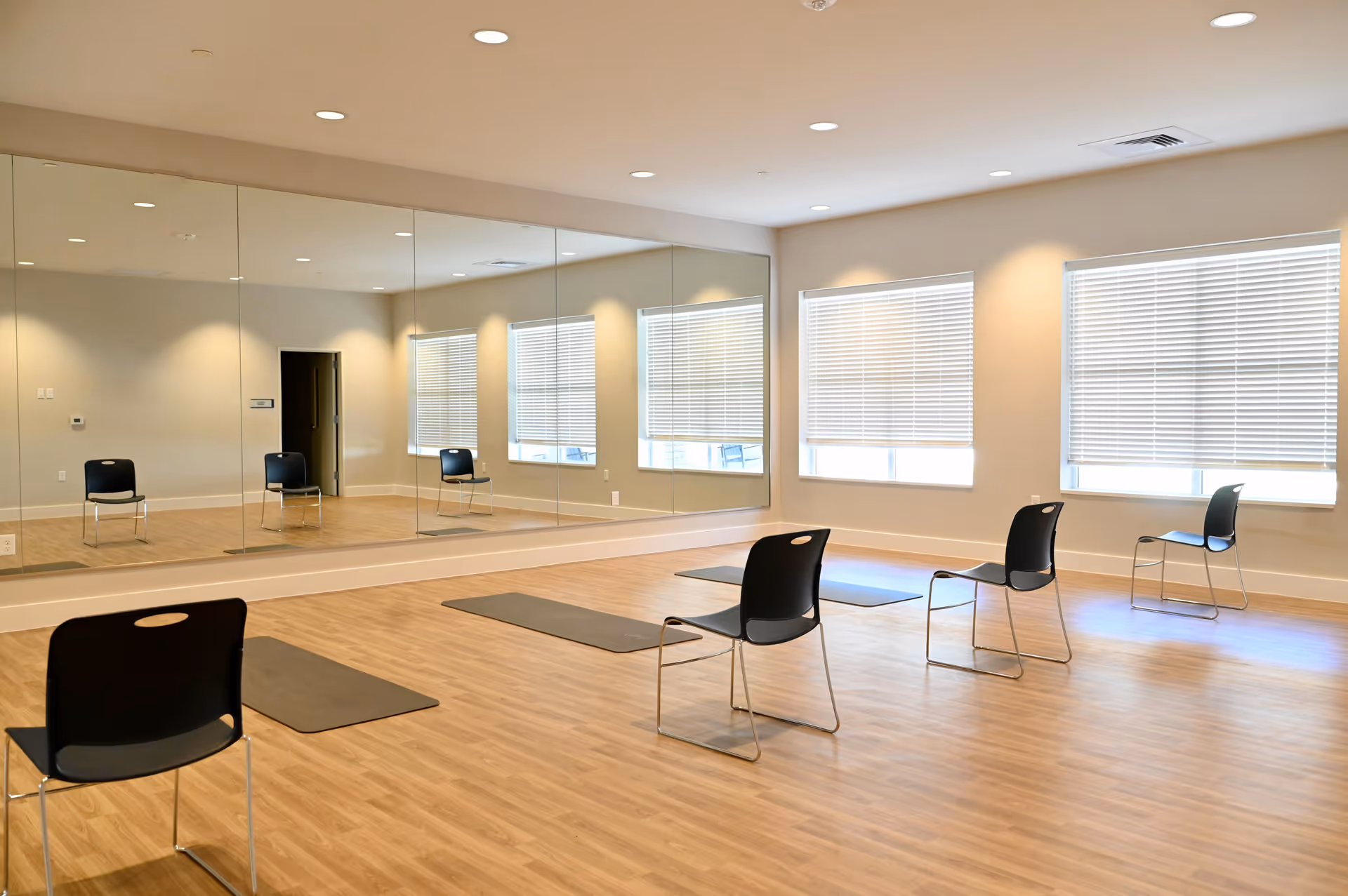 Bright multipurpose exercise studio with floor-to-ceiling mirrors, chairs and exercise mats on a wood floor.