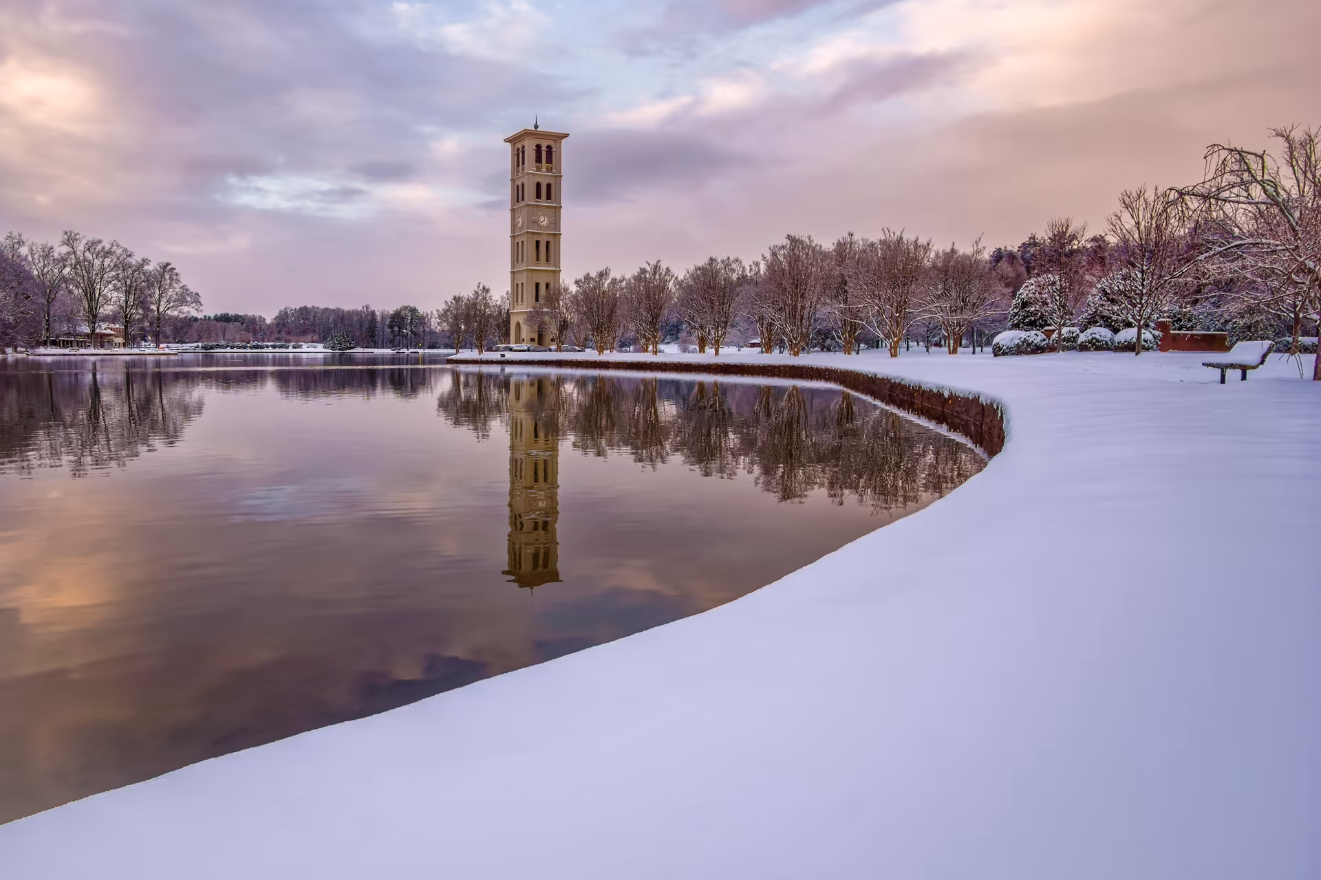 A serene winter landscape featuring a tall clock tower reflected in a calm lake, surrounded by snow-covered ground and leafless trees under a cloudy sky with soft pastel colors.