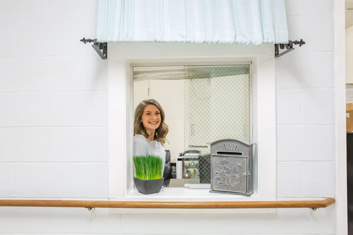 A smiling woman with shoulder-length hair sits behind a glass window in a white room. In front of the window, there is a small potted plant with green grass and a decorative metal mailbox labeled 'MAIL'. The window has a light blue fabric awning above it and a wooden handrail below.