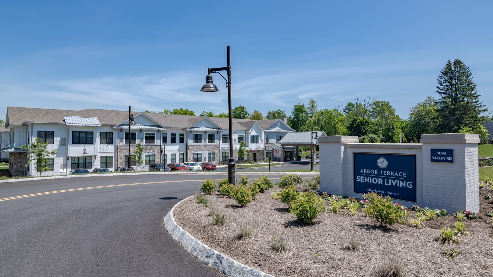 Exterior view of Arbor Terrace Basking Ridge senior living facility with a curved driveway, landscaped area with shrubs, and a large sign displaying the facility name and address. The building is two stories with white siding, stone accents, and multiple windows under a clear blue sky.