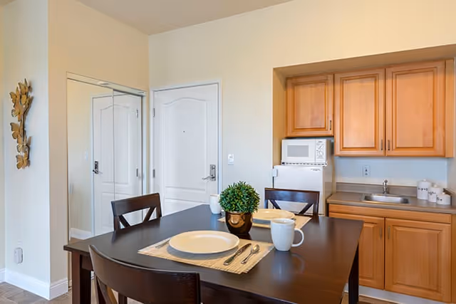 A small dining area with a dark wooden table set for two, including white plates, mugs, and silverware on beige placemats. Behind the table is a kitchenette with light wood cabinets, a small sink, a microwave, and a mini refrigerator. A white door and a mirrored closet door are visible in the background, along with a decorative wall piece on the left.