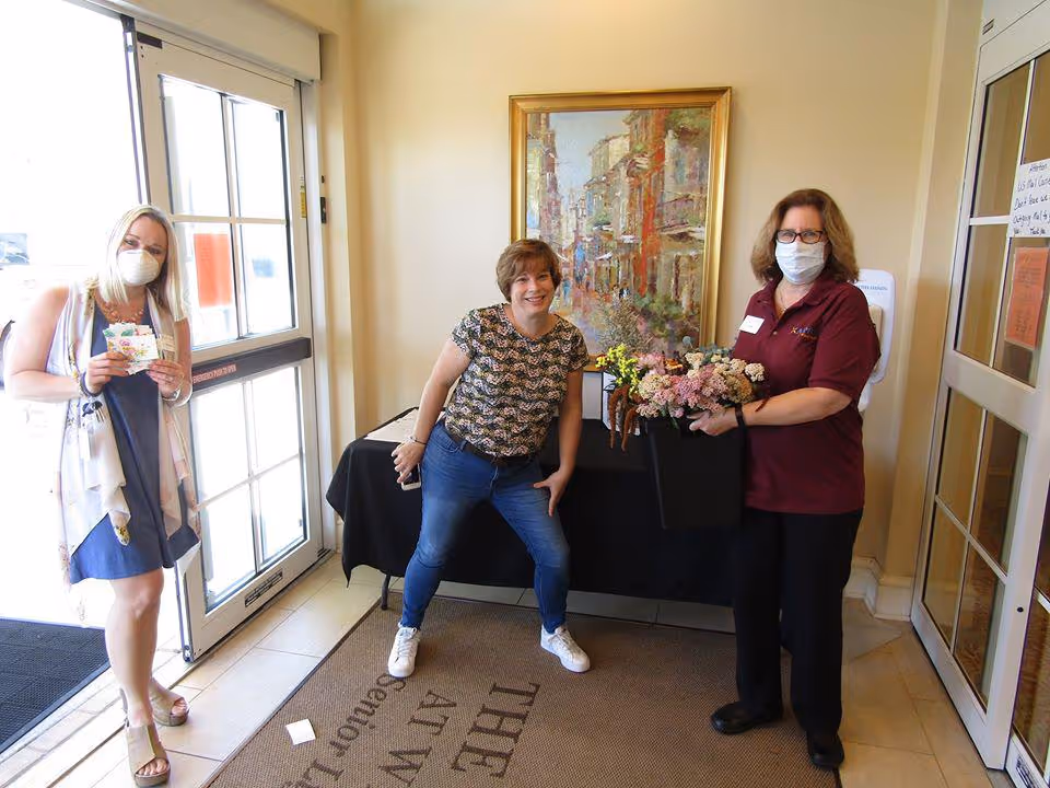 Three women standing inside the entrance of a facility. One woman on the left is wearing a mask and holding a card, the woman in the middle is smiling and posing with one hand on her hip, and the woman on the right is wearing a mask and holding a basket of flowers. There is a painting on the wall behind them and a table covered with a black cloth.