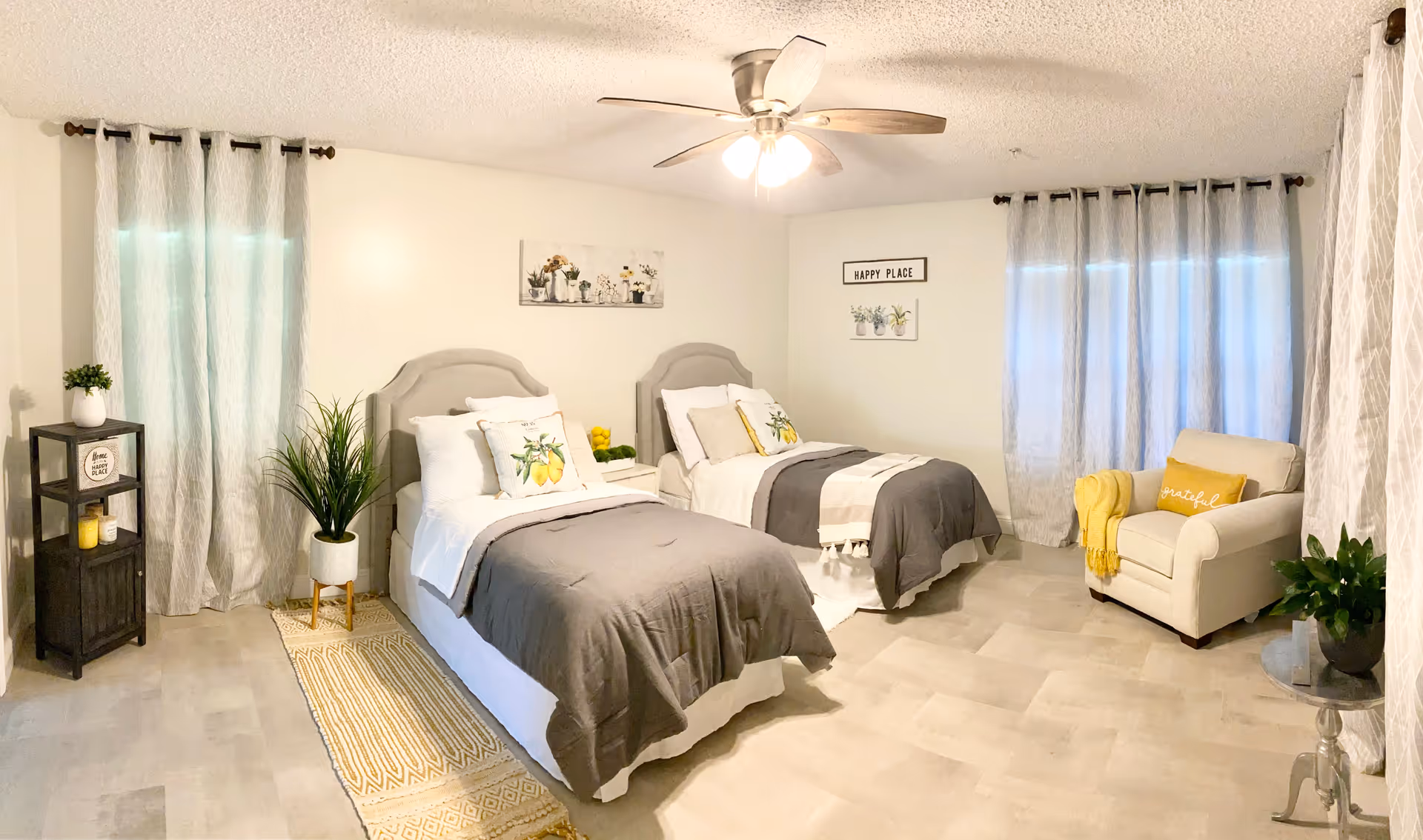 A bright and cozy bedroom with two twin beds, each with gray headboards, white bedding, and gray blankets. Decorative pillows with lemon designs are on the beds. The room has light-colored walls and floor tiles, two windows with sheer curtains, a ceiling fan with lights, a beige armchair with a yellow throw blanket and pillow, and several plants and wall art including a sign that says 'HAPPY PLACE'.