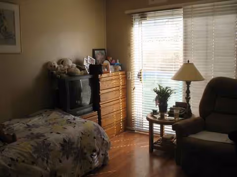 Cozy bedroom with a bed, dresser, TV, window blinds, and a side table with a lamp.