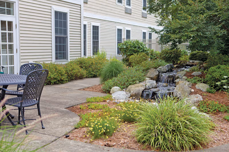 Outdoor patio area with a metal table and chairs next to a building with beige siding and multiple windows. A concrete pathway curves around a landscaped garden featuring green shrubs, yellow flowers, rocks, and a small cascading water feature.