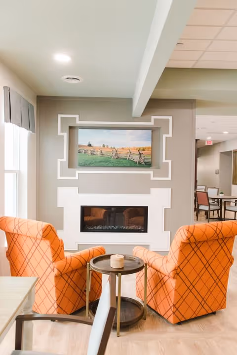 Two orange plaid armchairs face a built-in fireplace with a landscape painting above, separated by a small round table in a bright common area.