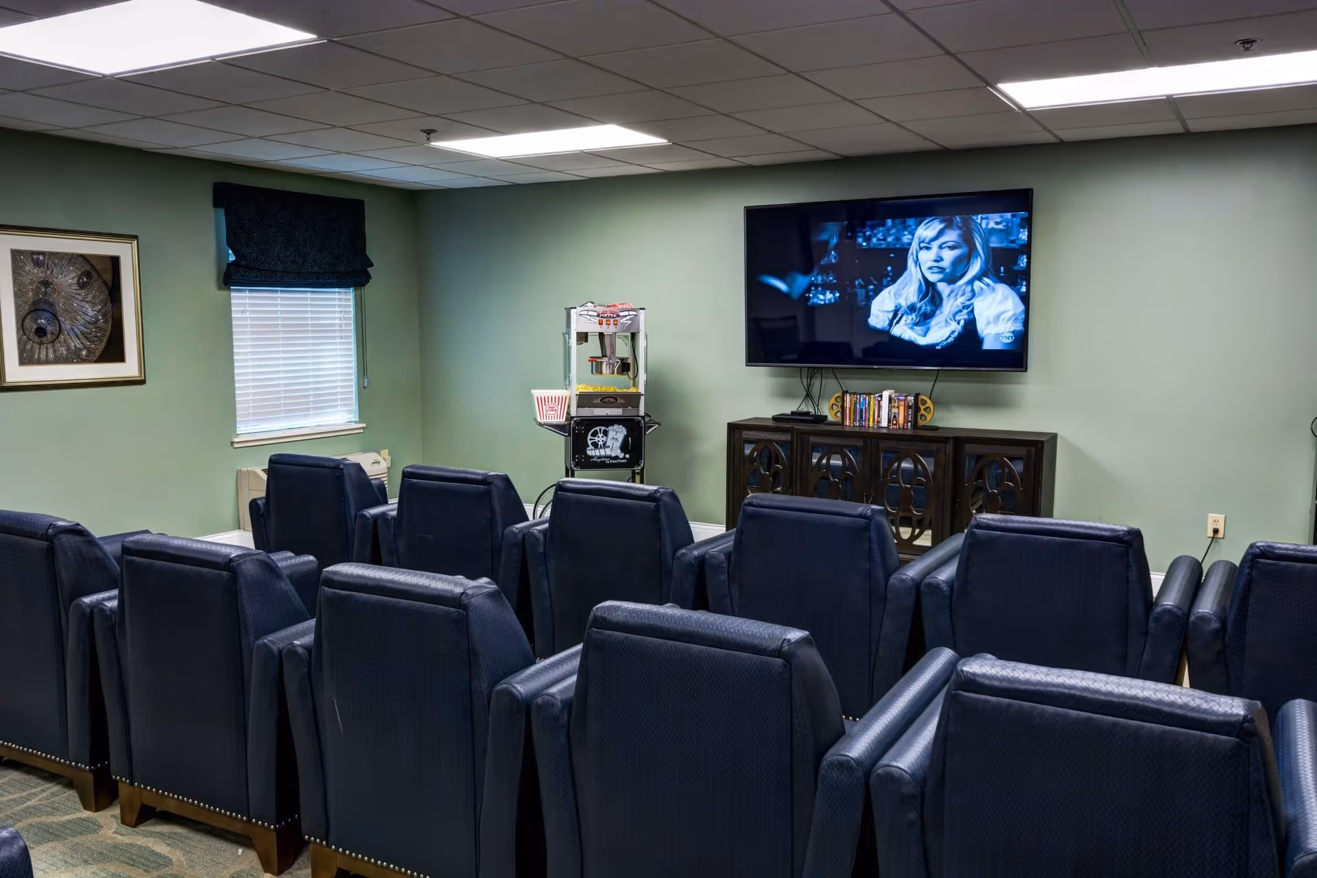 A small movie theater room with two rows of dark blue cushioned chairs facing a wall-mounted flat screen TV playing a black and white movie. There is a popcorn machine and a small cabinet with books and DVDs below the TV. The walls are painted light green and there is a window with a dark blue valance on the left side.