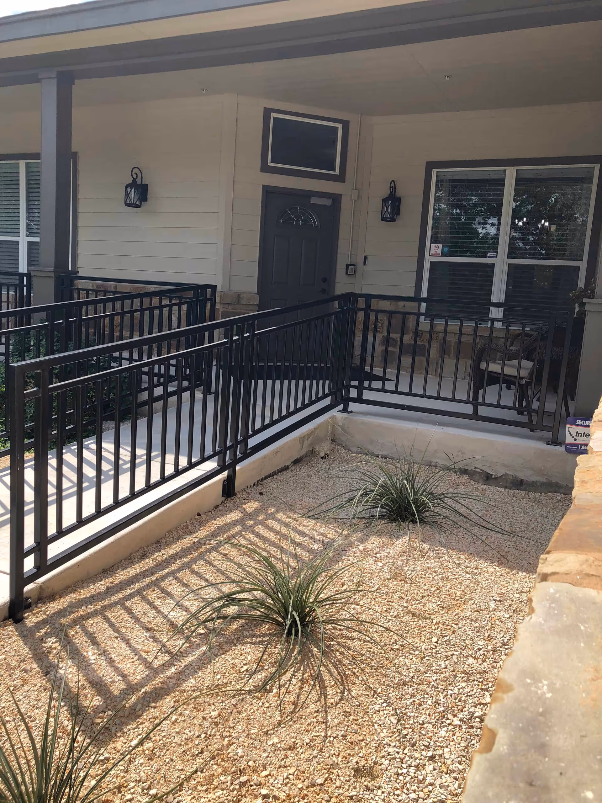 Outdoor view of a building entrance with a black metal ramp railing leading to a dark-colored door. There are two wall-mounted lantern-style lights on either side of the door, a window with blinds to the right, and a small gravel garden area with some plants in front of the ramp.