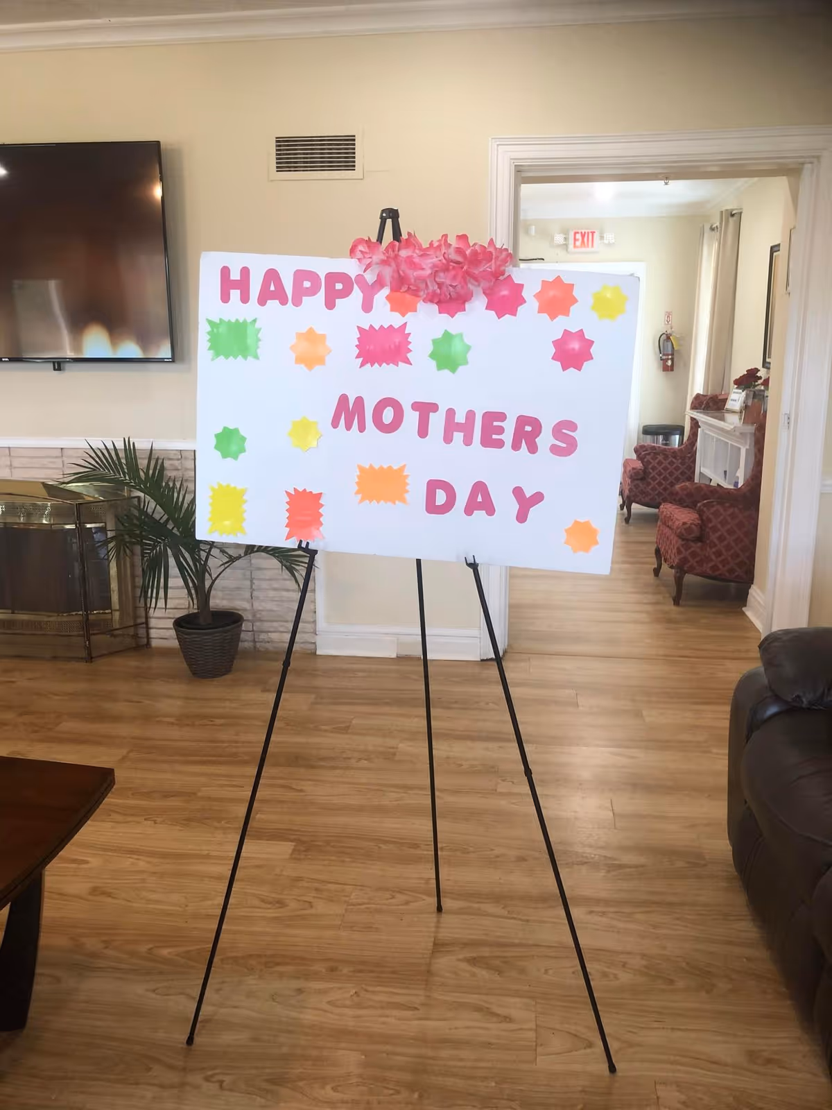A decorated sign on an easel that reads "HAPPY MOTHERS DAY" with colorful paper stars and flowers in a senior living facility common room.