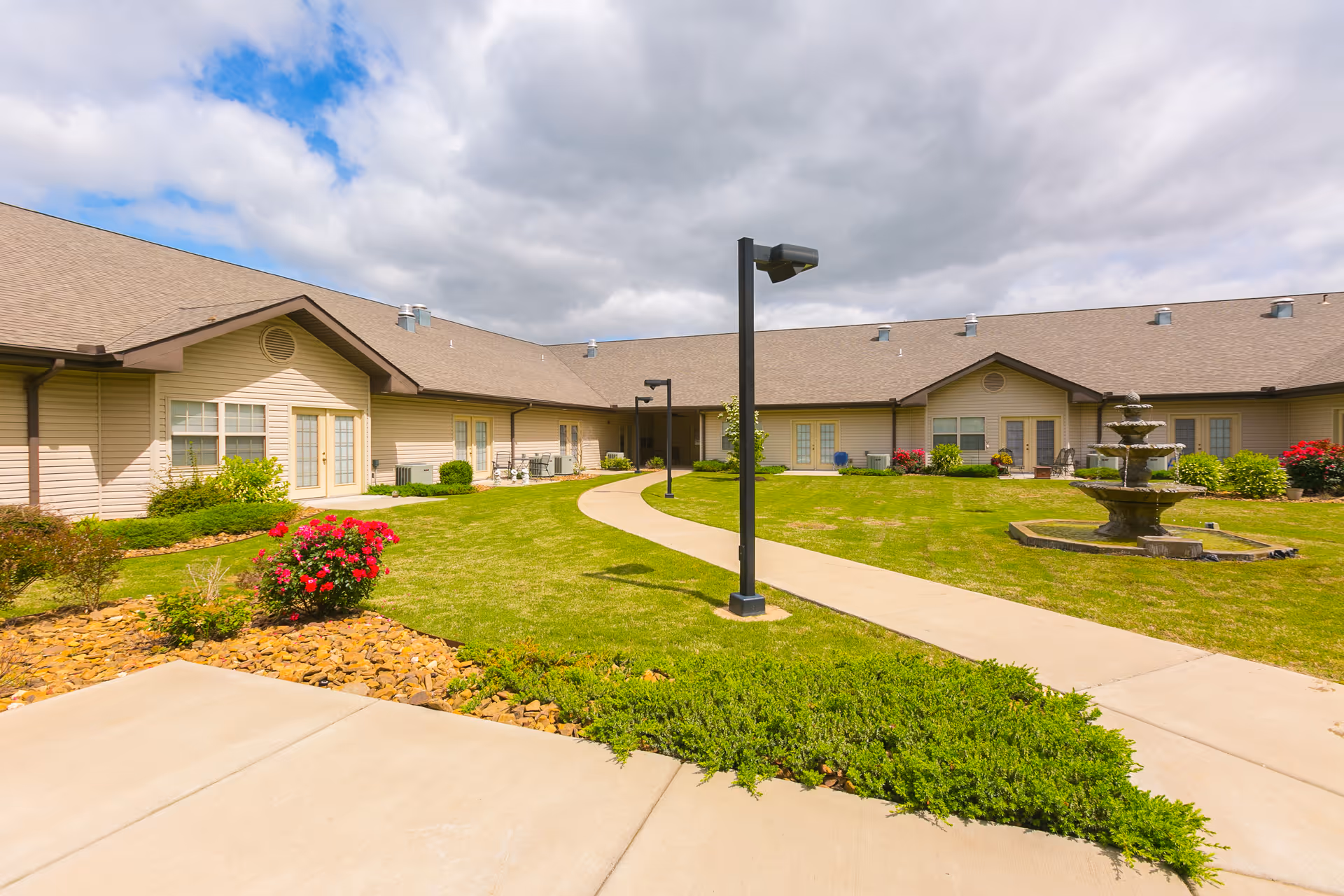 Outdoor courtyard area of The Bungalows at Fayetteville featuring a paved walkway, green lawn, flower beds with red and pink flowers, a multi-tiered water fountain, and beige single-story buildings with multiple windows and doors under a partly cloudy sky.