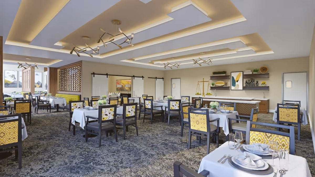 A spacious, well-lit dining room with multiple tables covered in white tablecloths, each set with plates, glasses, and silverware. The chairs have patterned yellow and gray upholstery. The ceiling features modern recessed lighting and decorative fixtures. The room has a carpeted floor and neutral-colored walls with shelves and artwork.