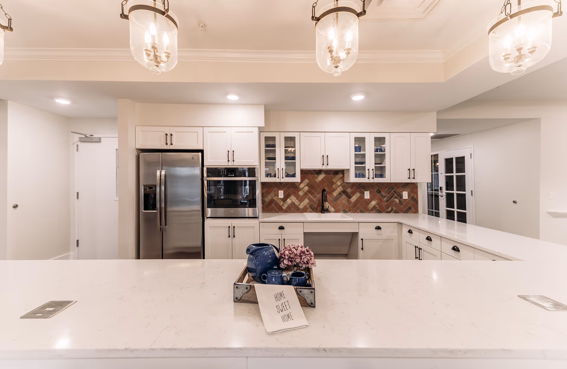 Modern kitchen with white cabinetry, stainless steel refrigerator, built-in oven, and a brick-patterned backsplash. A large white marble countertop island is in the foreground with a decorative tray holding a blue teapot set and a cloth that reads 'HOME SWEET HOME'. Three pendant lights hang from the ceiling.