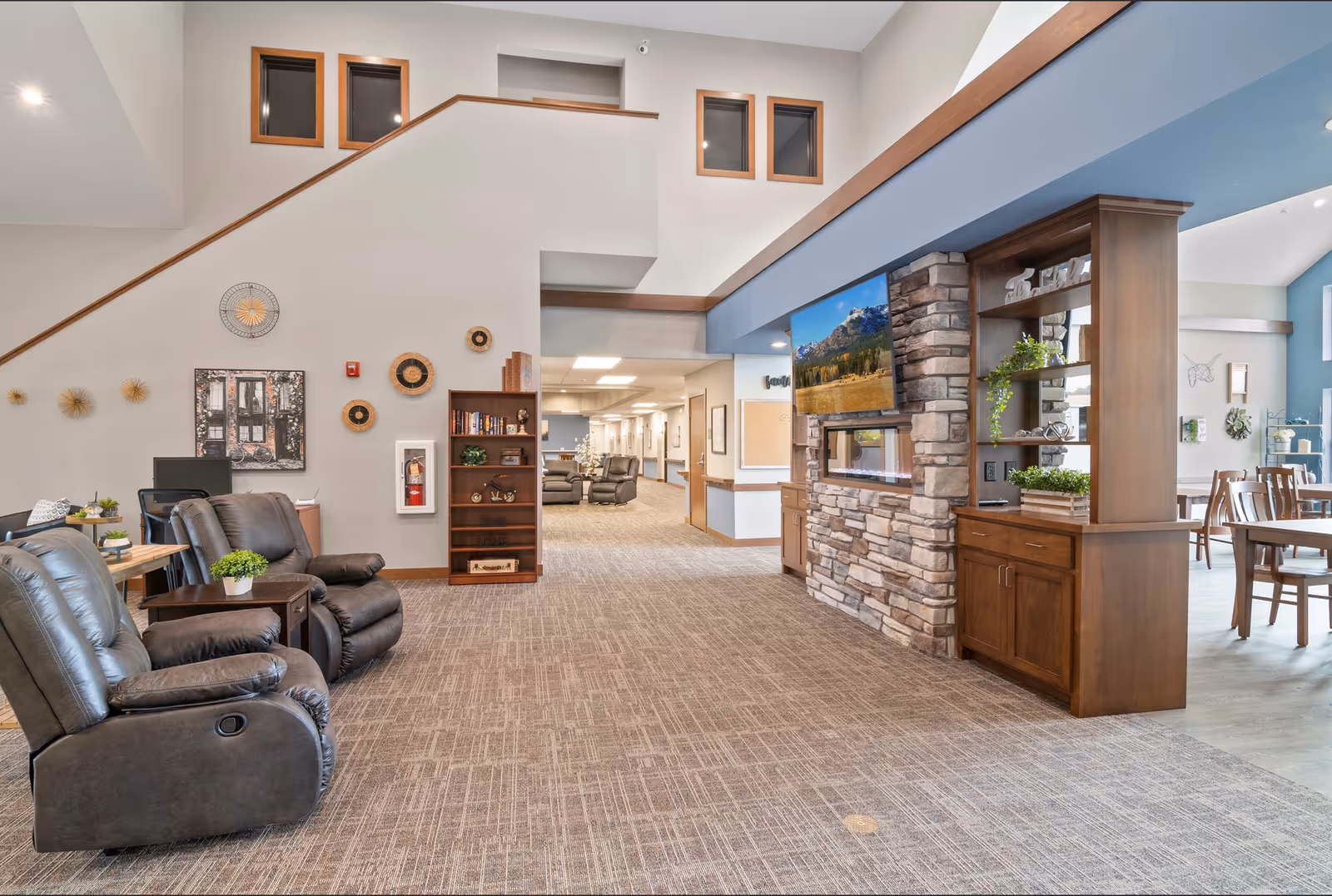 A spacious and well-lit common area in a senior living facility featuring comfortable leather recliners, a stone fireplace with a mounted flat-screen TV above it, wooden shelving with decorative plants, and a dining area with wooden tables and chairs in the background. The walls are decorated with art and woven wall hangings, and there is a staircase leading to an upper level.