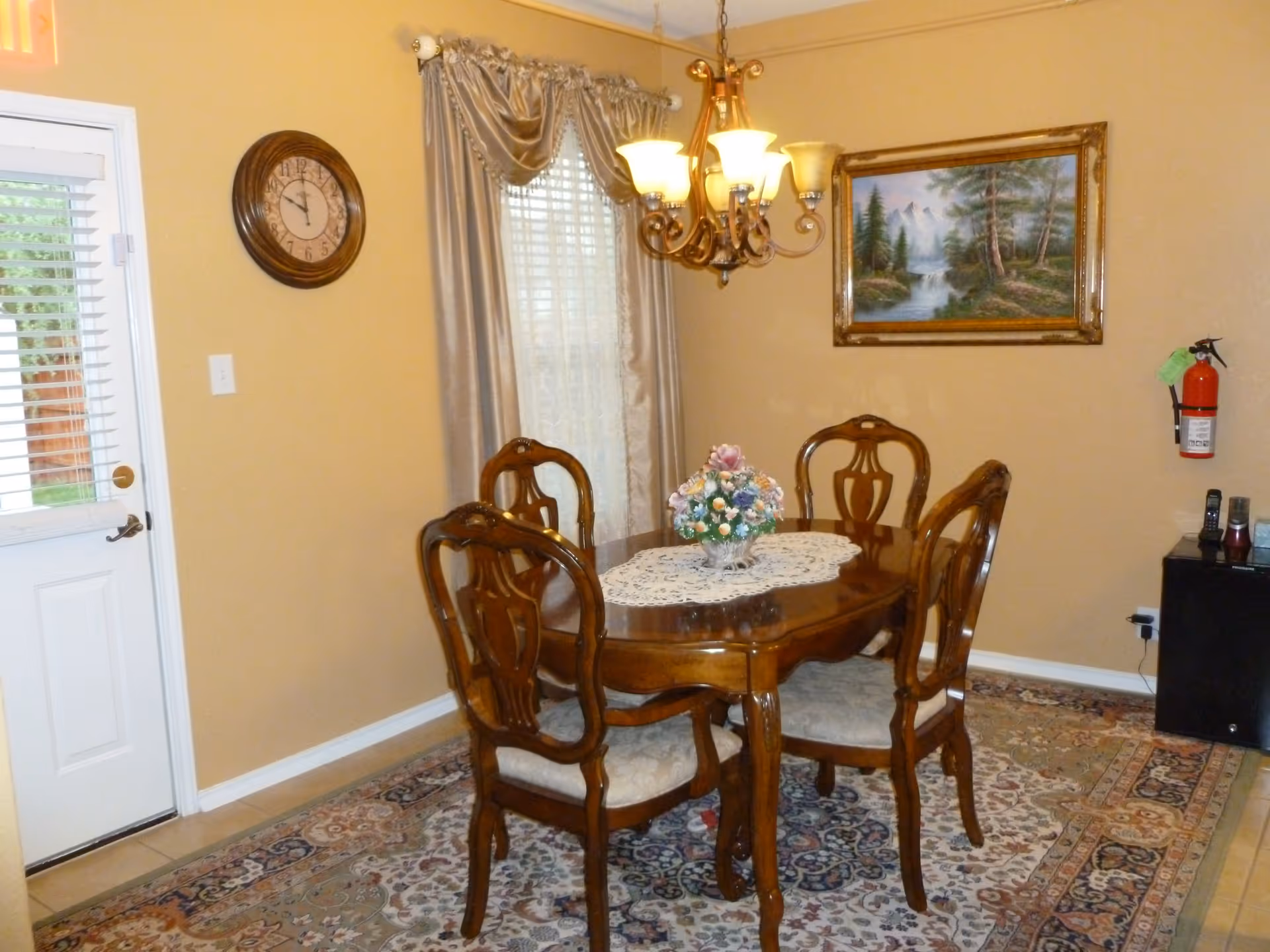 A dining room with a wooden dining table and four matching chairs. The table has a lace doily and a floral centerpiece. There is a chandelier hanging above the table, a wall clock, a window with curtains, a framed landscape painting, and a fire extinguisher mounted on the wall. A door with glass panels is visible on the left side.