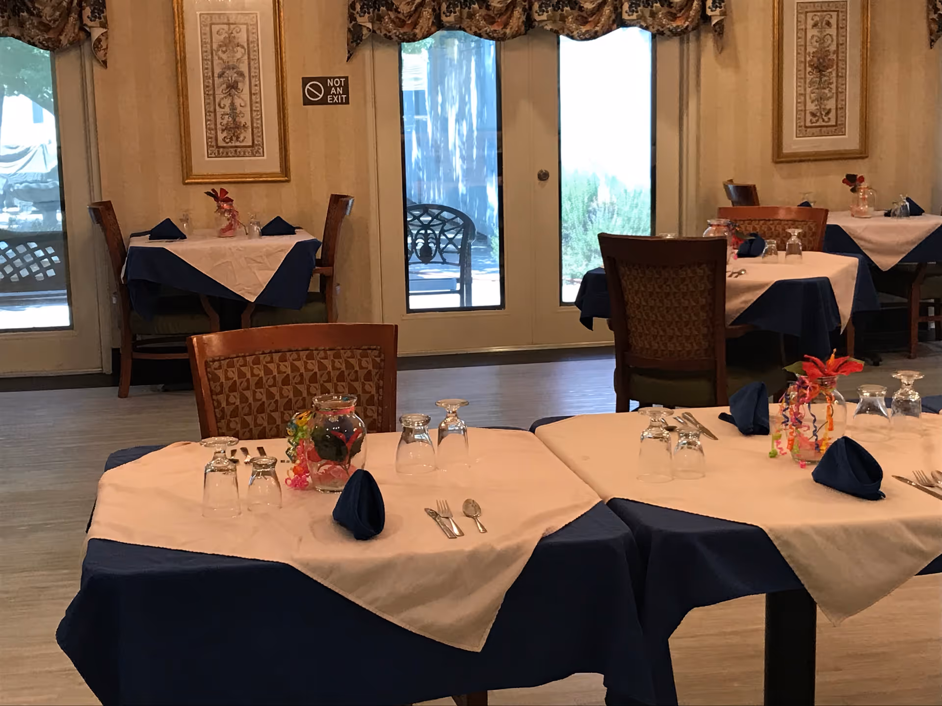 Dining room with tables set with blue and white tablecloths, folded napkins, glassware, and chairs in front of glass doors.
