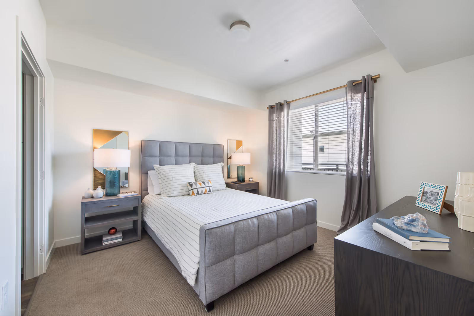 A bright bedroom with a gray upholstered bed featuring white and striped bedding. There are two matching nightstands with lamps and decorative items on either side of the bed. A window with gray curtains lets in natural light. A dark wooden dresser holds books, a framed photo, and decorative objects.