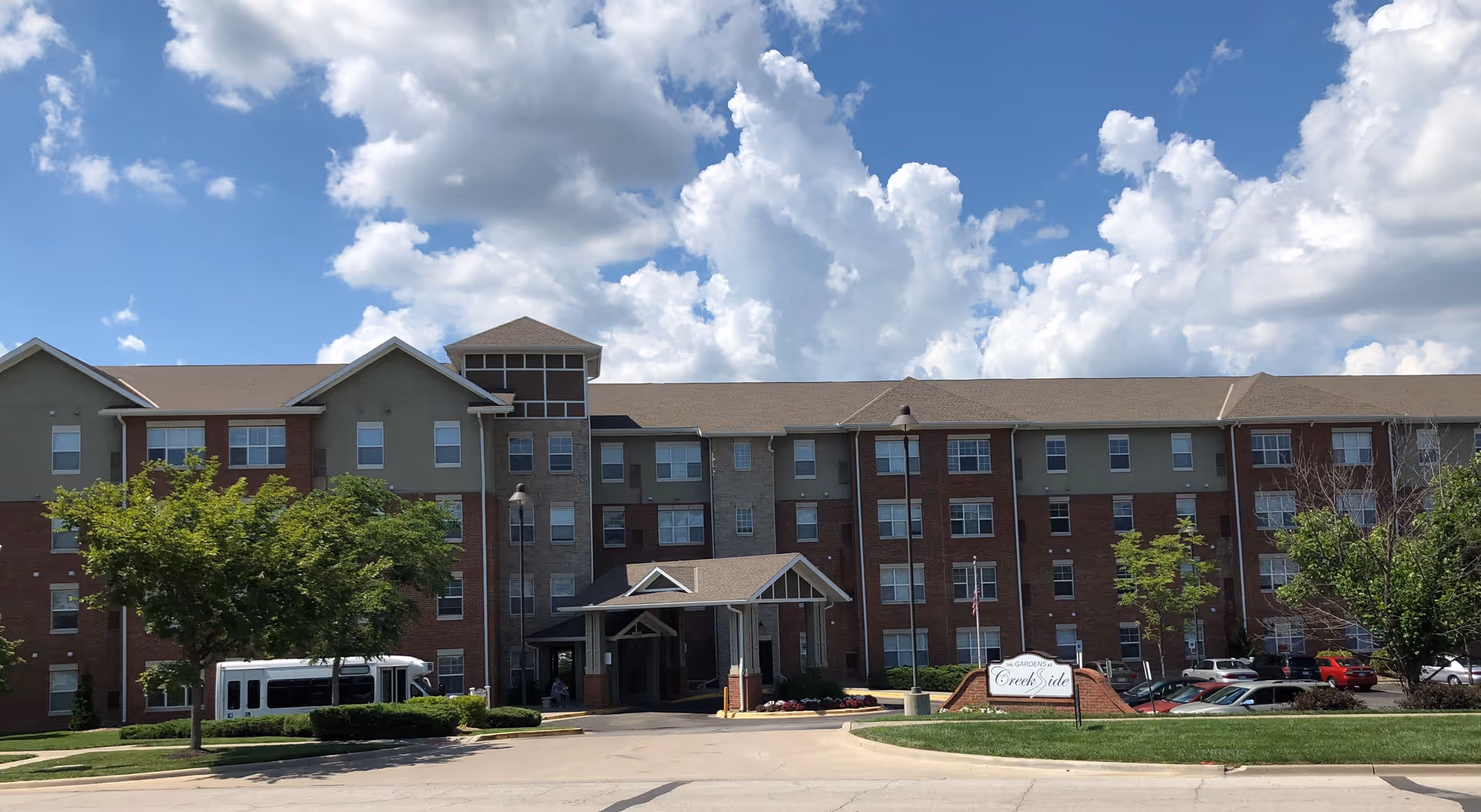 Front exterior view of a multi-story senior living facility building named The Gardens at Creekside, with a covered entrance, several windows, trees, parked cars, and a partly cloudy sky.