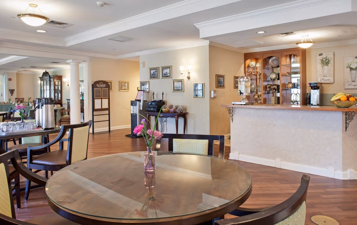 A dining area in a senior living facility with round tables and chairs. One table in the foreground has a glass top with a small vase holding pink flowers. In the background, there is a counter with a beverage dispenser, a coffee machine, and a bowl of fruit. The walls are decorated with framed pictures and floral artwork. The floor is wooden, and the lighting is warm with ceiling fixtures and wall sconces.