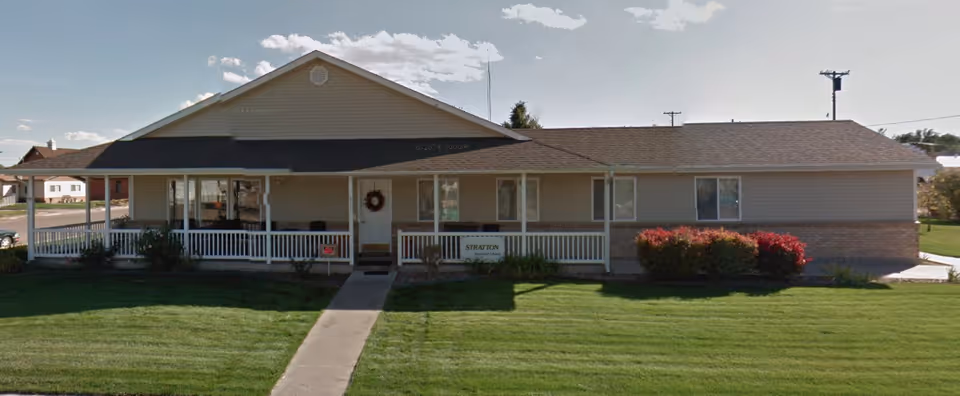 Single-story building with beige siding and a dark roof, featuring a covered front porch with white railings and a concrete walkway leading to the entrance. The lawn is neatly mowed, and there are bushes with red and green foliage near the porch.