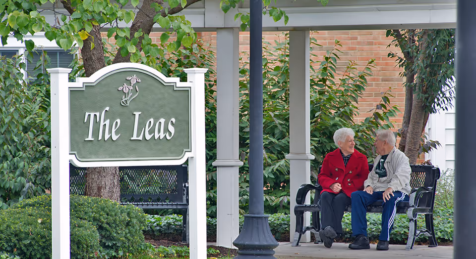 Two elderly people sitting on a black bench under a covered area, engaged in conversation. There is a green sign with white trim that reads 'The Leas' in the foreground, surrounded by greenery and trees.