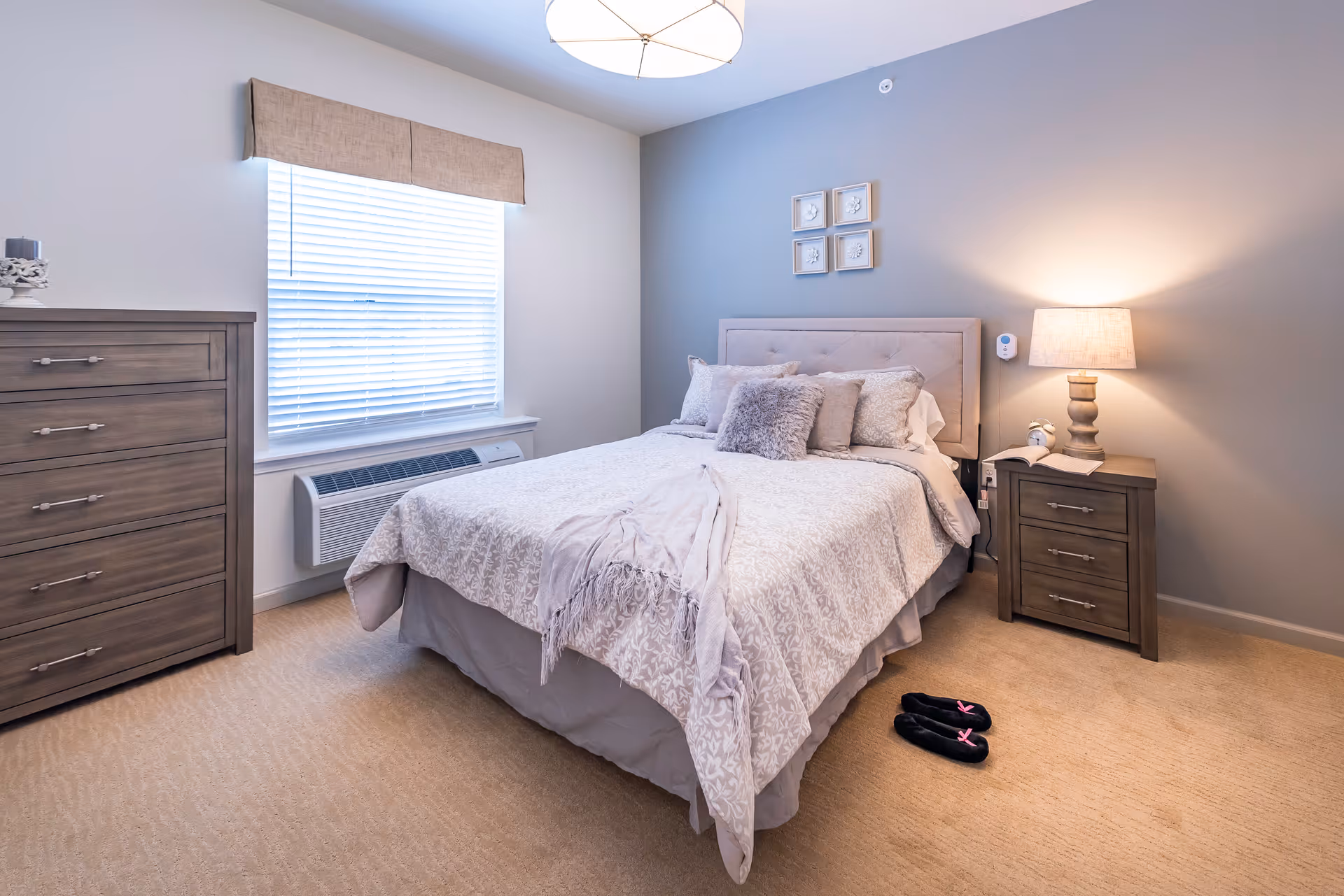 A neatly made bed with light-colored bedding and multiple pillows in a bedroom. There is a wooden dresser to the left and a matching nightstand with a lamp on the right. A window with blinds and a beige valance is on the left wall. A pair of black slippers with pink bows is on the carpeted floor near the bed. The walls are painted in light gray tones, and there are four small framed artworks above the headboard.