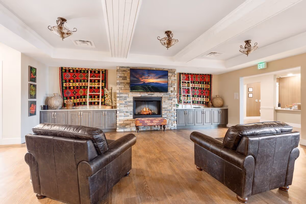 A cozy living room area with two dark brown leather armchairs facing a stone fireplace with a mounted flat-screen TV above it. On either side of the fireplace are gray cabinets topped with decorative items and colorful patterned wall hangings. The room has wooden flooring, white ceiling beams, and warm lighting. An exit sign is visible above a doorway leading to another room.