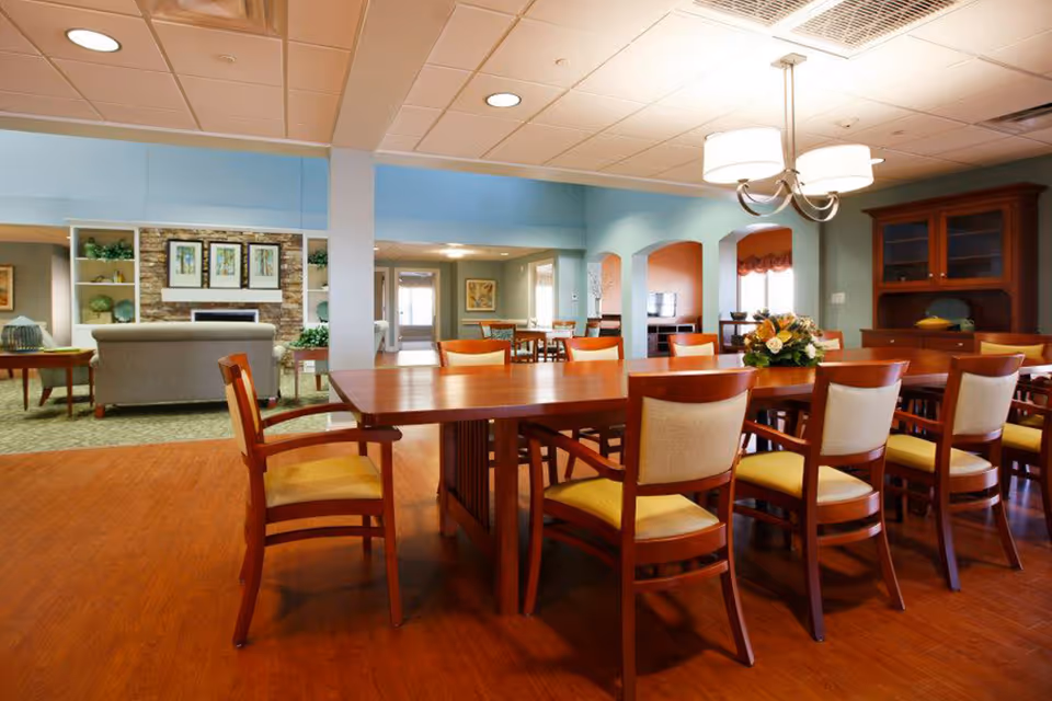 A spacious dining area in a senior living facility with a long wooden table surrounded by wooden chairs with cushioned seats. The room features a chandelier overhead, a wooden cabinet, and an adjacent living room area with sofas and framed artwork on the walls.