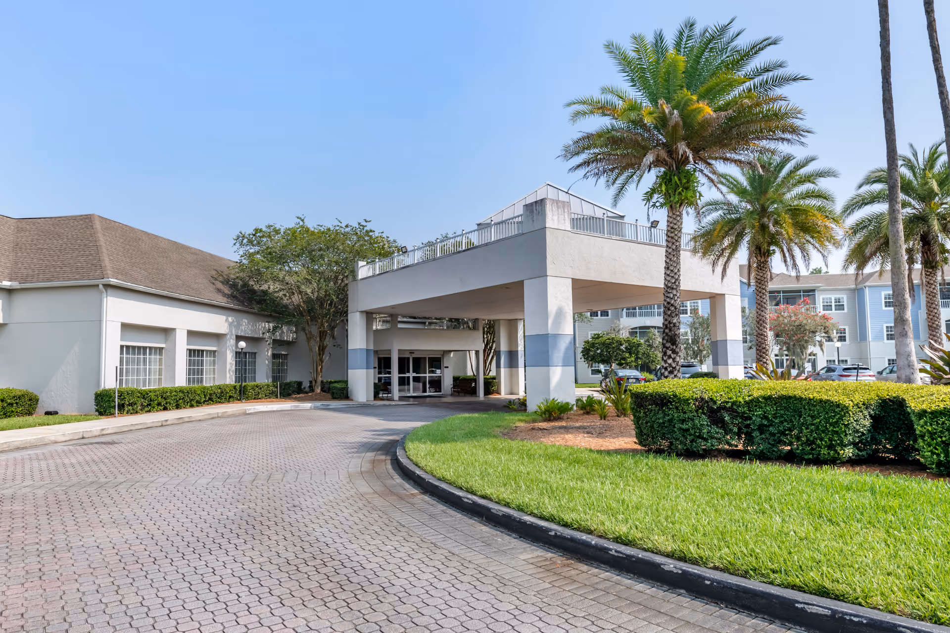 Exterior view of Brookdale Southside facility entrance with a covered drop-off area, palm trees, manicured bushes, and a paved driveway under a clear blue sky.