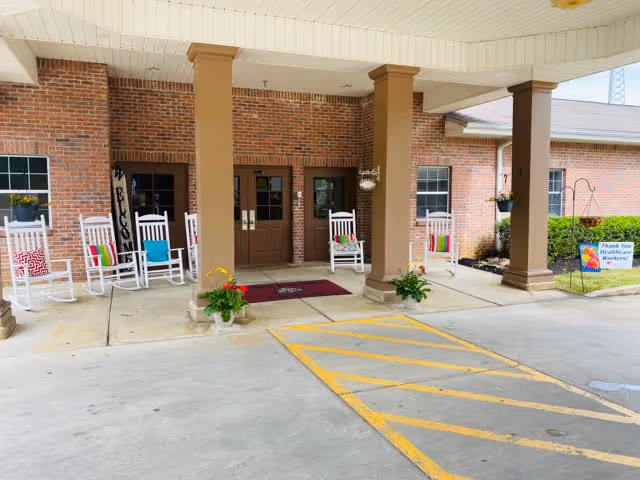 Covered brick entrance with columns, white rocking chairs, potted plants, and a drive-up drop-off area.