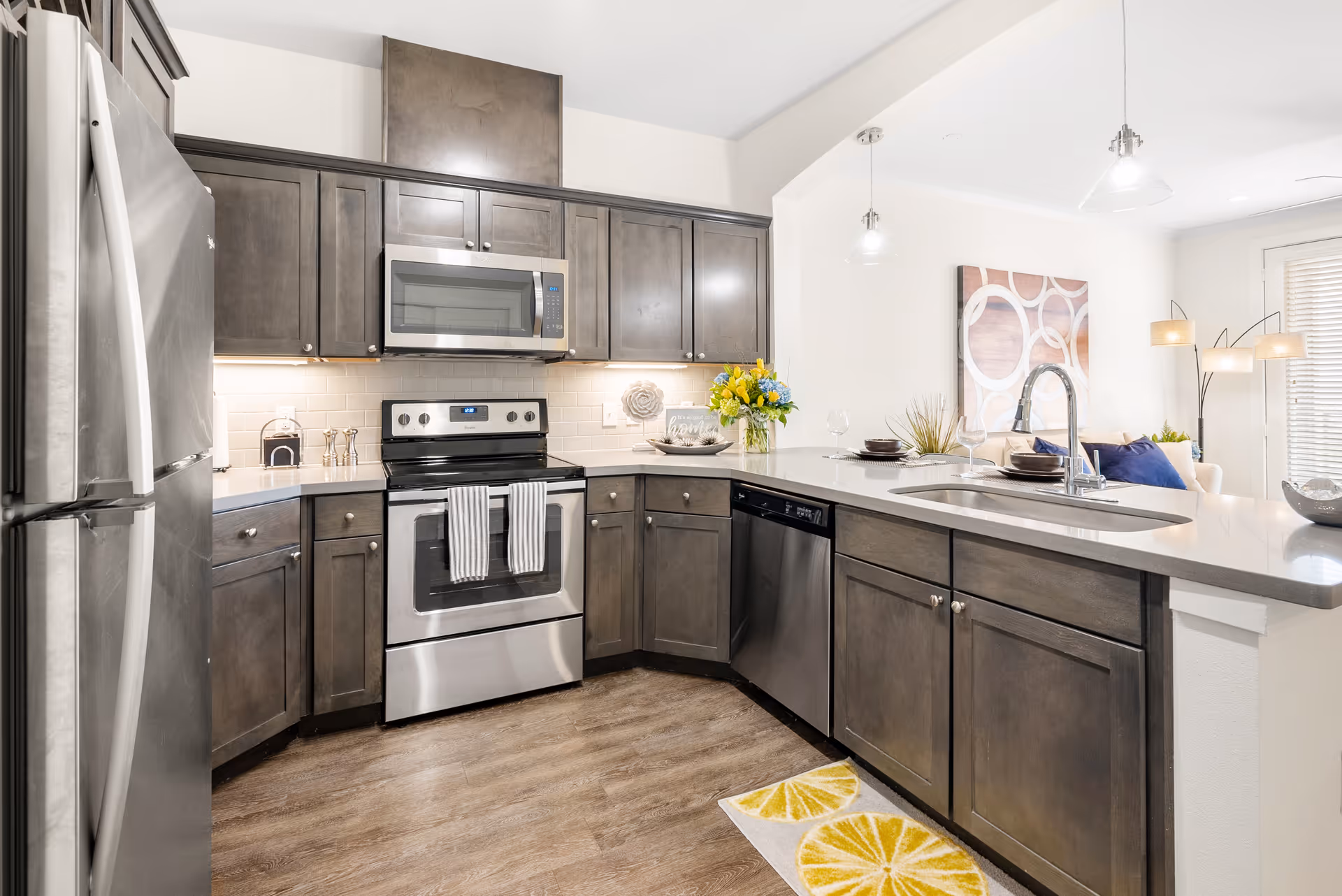 Modern kitchen with stainless steel appliances including a refrigerator, microwave, stove, and dishwasher. Dark wood cabinets with silver knobs, white countertops, and a sink with a high-arc faucet. The kitchen opens to a living area with a sofa, floor lamp, and wall art. The floor is wood, and there is a yellow and white lemon-patterned rug in front of the sink.