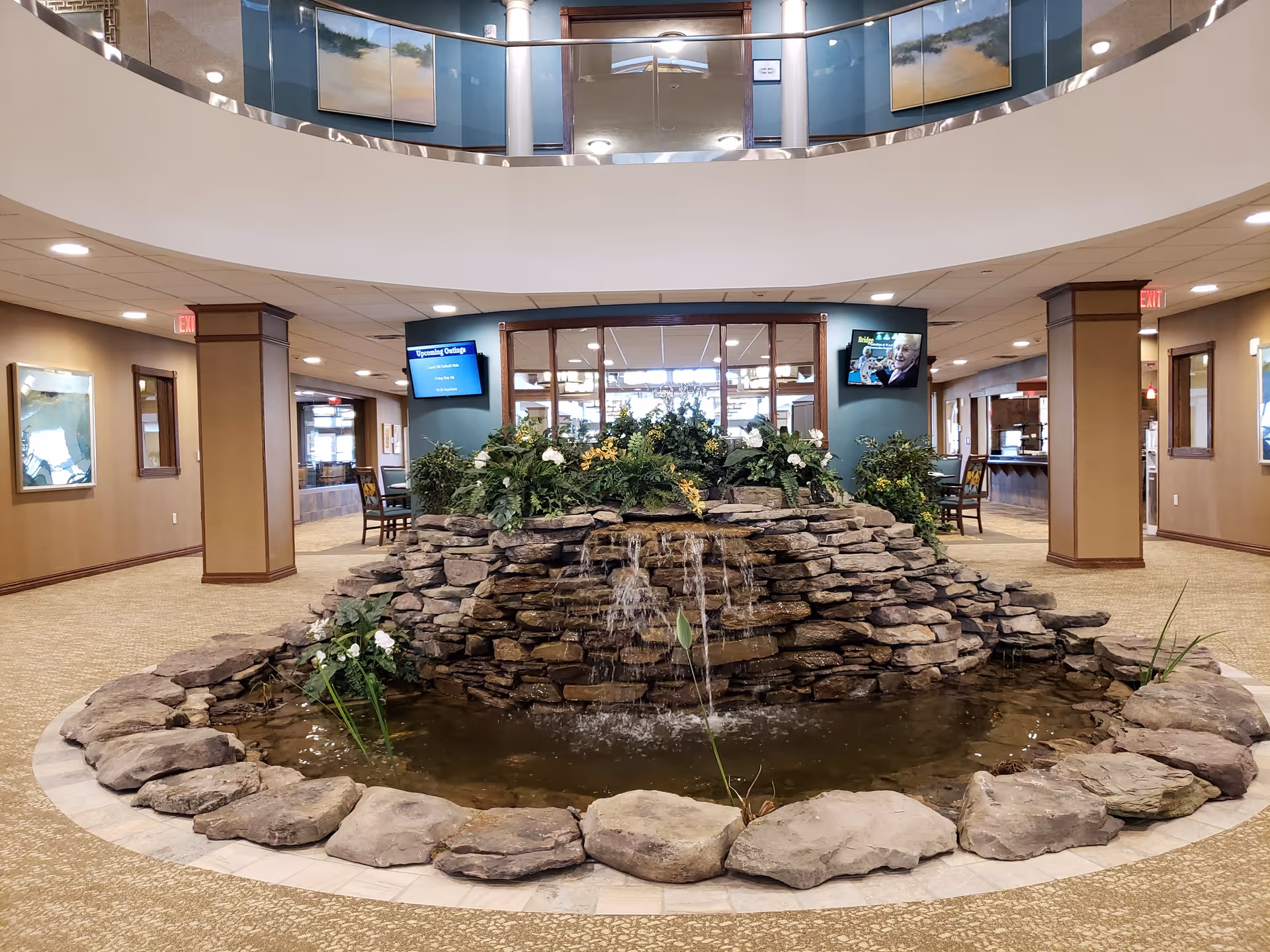 Indoor water feature with a stone waterfall surrounded by plants in the center of a spacious lobby area with carpeted floors, columns, and seating areas. The lobby has a second-floor balcony with glass railing and paintings on the walls.