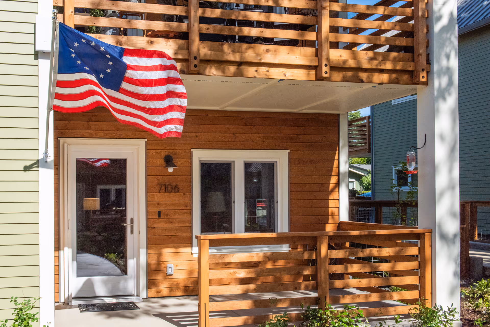 Exterior view of a senior living community unit with wooden siding and a small porch. An American flag is mounted on the left side near the entrance door, and there is a balcony above the porch. The unit number 7106 is visible next to the door.
