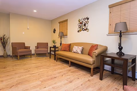 A cozy living room with a tan sofa adorned with three decorative pillows, two brown armchairs, two dark wooden side tables each with a lamp, a wall decoration above the sofa, wooden blinds on the windows, and a light wood floor.