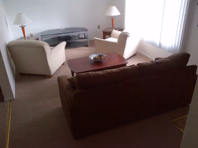 Living room with a brown sofa, two cream armchairs, a wooden coffee table, TV stand and floor lamps by a bright window.