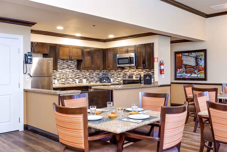 Dining area with set tables and chairs in front of an open kitchen with dark wood cabinets, stainless steel appliances, and a mosaic tile backsplash.