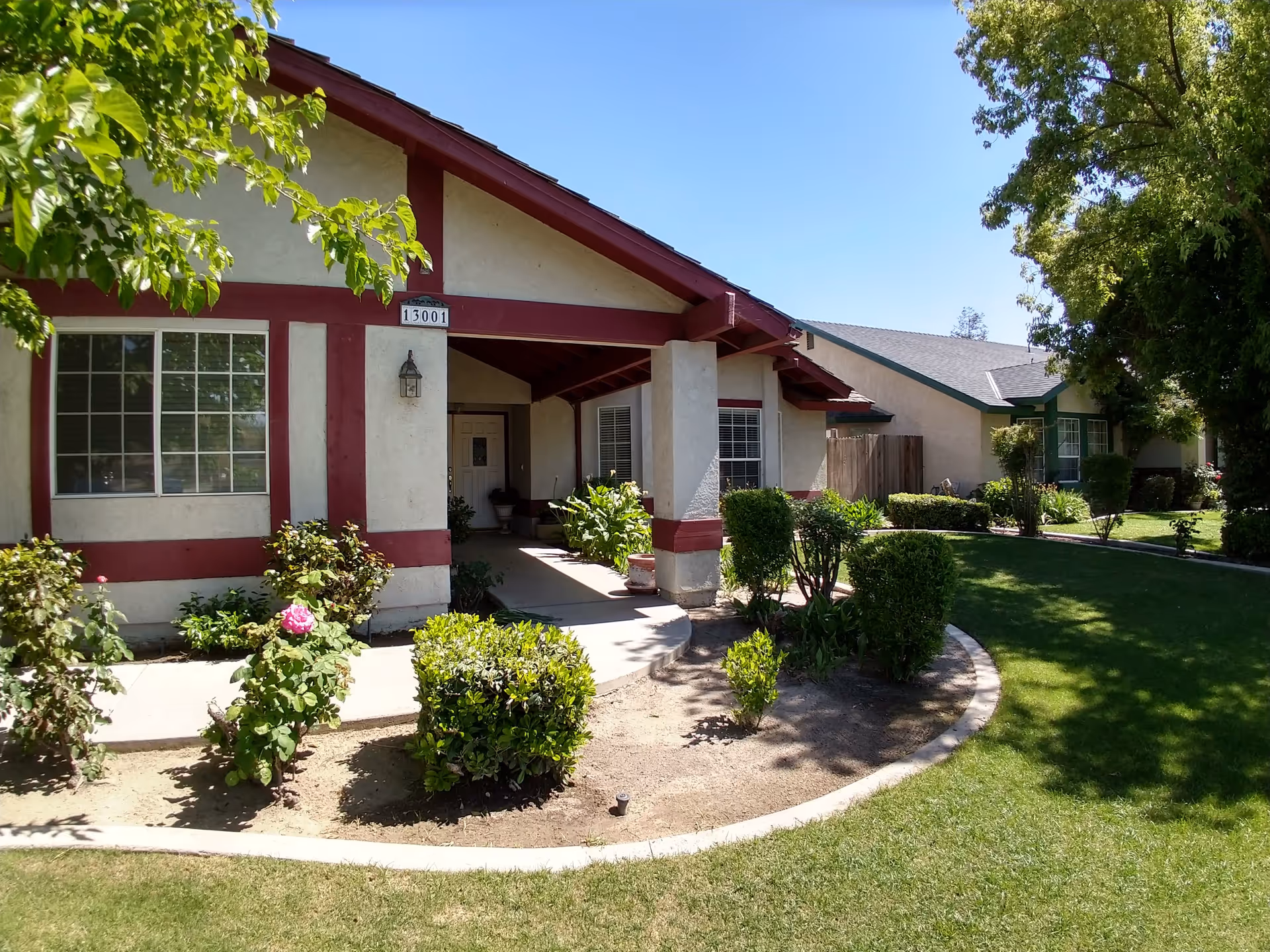 Exterior view of a single-story residential building with beige walls and red trim. The building has a covered porch entrance with a door and windows. There are well-maintained bushes, plants, and a lawn in front of the building under a clear blue sky.
