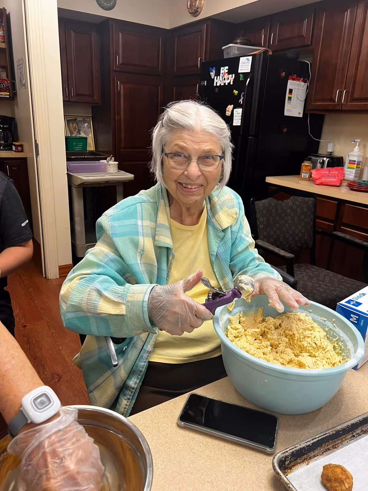 An elderly woman wearing glasses and a plaid jacket is smiling while scooping dough from a large blue bowl in a kitchen. She is wearing disposable gloves and sitting at a counter with a smartphone and a baking tray with a single cookie. The kitchen has dark wooden cabinets and a black refrigerator in the background.