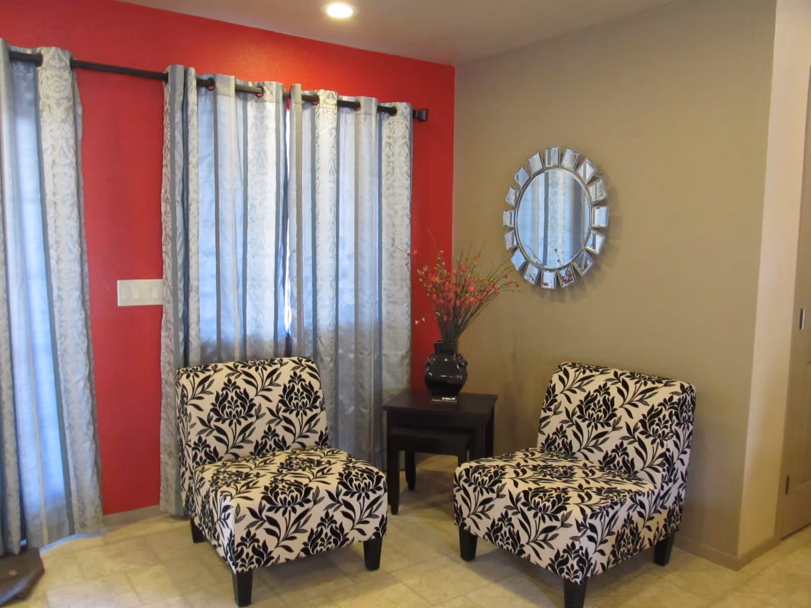 A cozy corner of a living room with two black and white patterned chairs facing each other, a small dark wooden side table between them holding a black vase with pink flowers, a round decorative mirror on the beige wall, and light blue curtains covering windows on a red accent wall.
