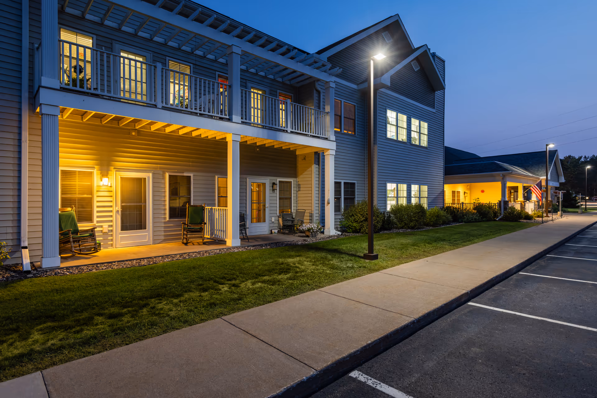 Exterior view of Milestone Senior Living Woodruff at dusk, showing a two-story building with lit windows, a covered porch with rocking chairs, a well-maintained lawn, sidewalk, and a parking lot with street lamps.