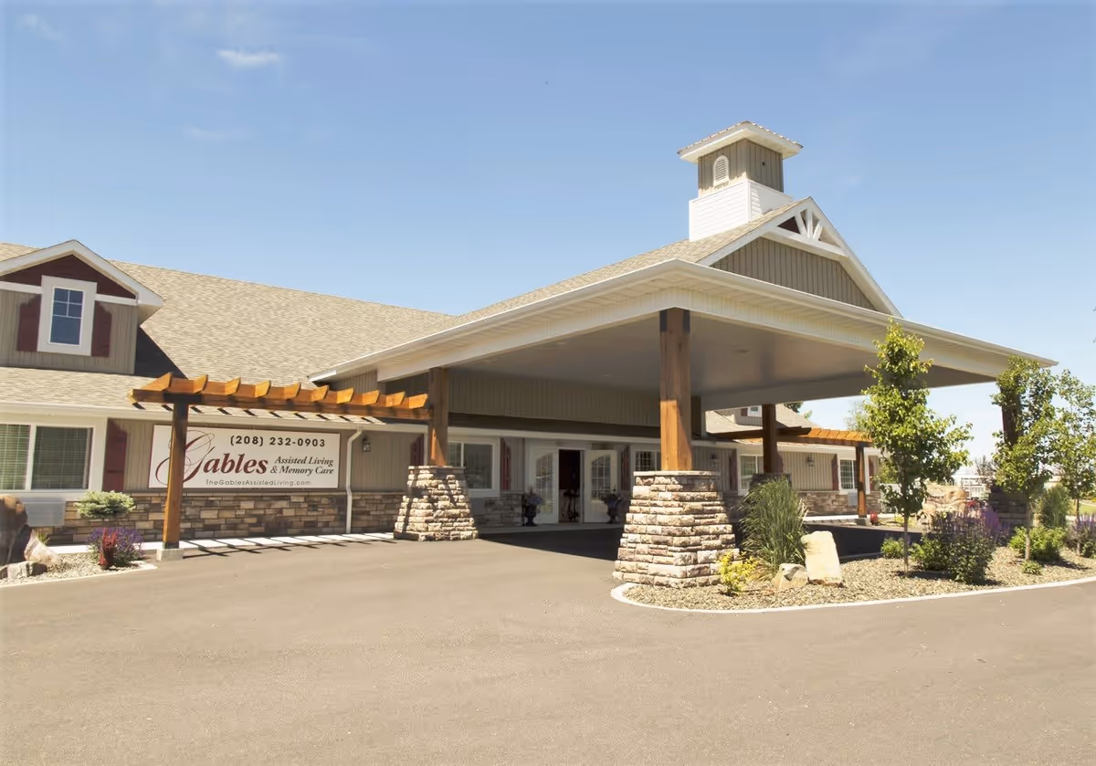 Exterior view of The Gables Memory Care of Pocatello building with a covered entrance supported by stone and wooden pillars, a pergola, and landscaped greenery under a clear blue sky.