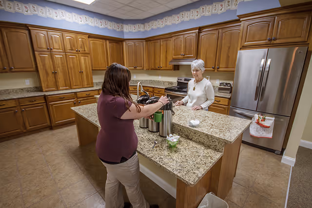 Two women stand at a granite island in a spacious kitchen with wooden cabinets and a stainless steel refrigerator.