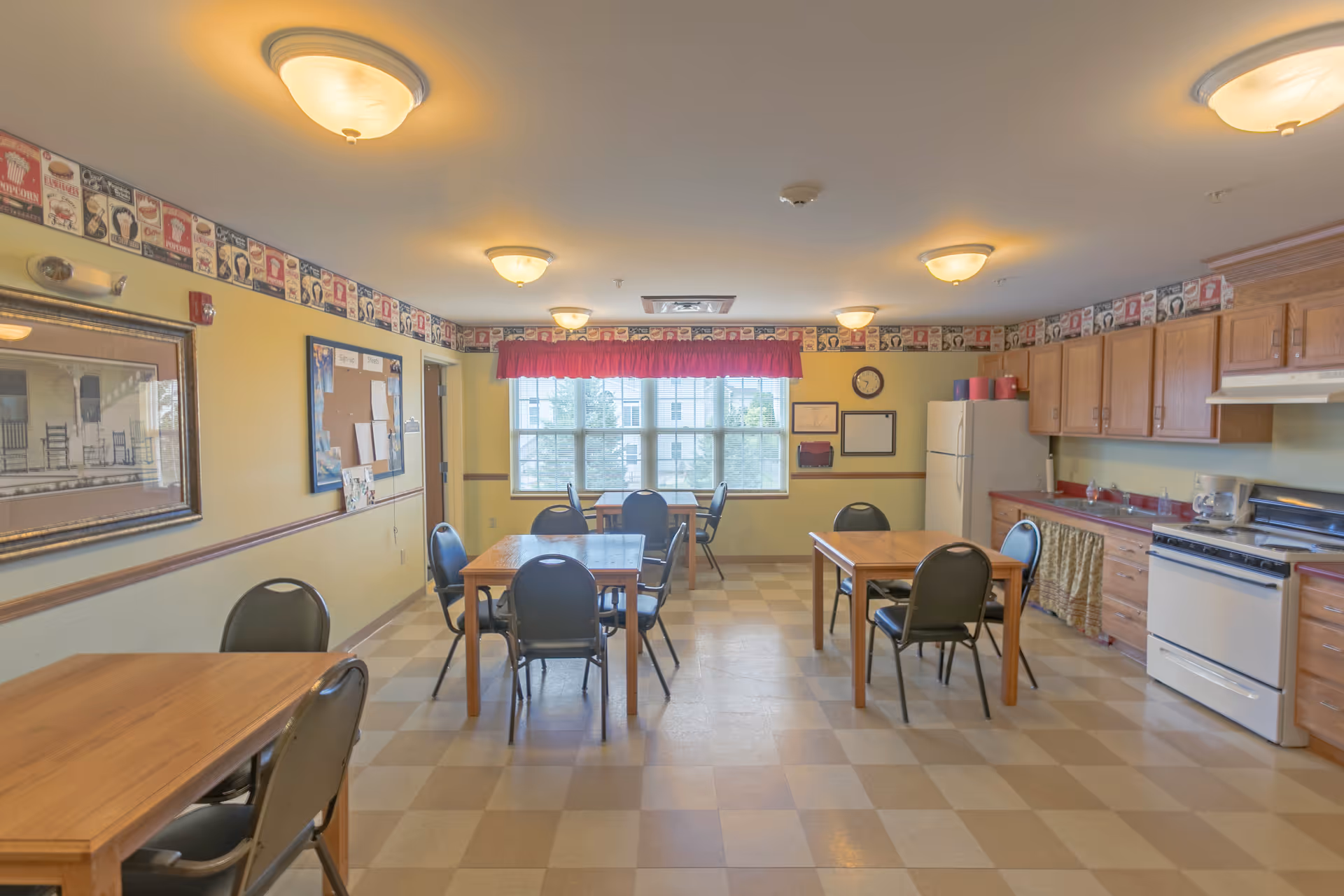 A communal dining area with several wooden tables and black chairs. The room has a checkered floor, yellow walls with decorative border near the ceiling, and multiple ceiling lights. There is a kitchen area on the right side with wooden cabinets, a white refrigerator, a stove, and a coffee maker. A large window with a red valance lets in natural light at the far end of the room.